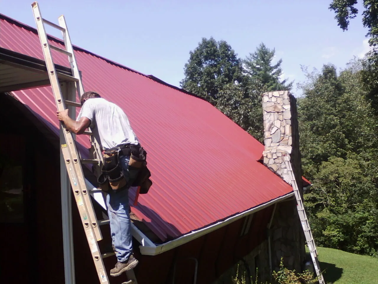 Roofer on a ladder beside a red metal roof and stone chimney — Soffit & Fascia Repair by Roofing Brothers in Craigville
